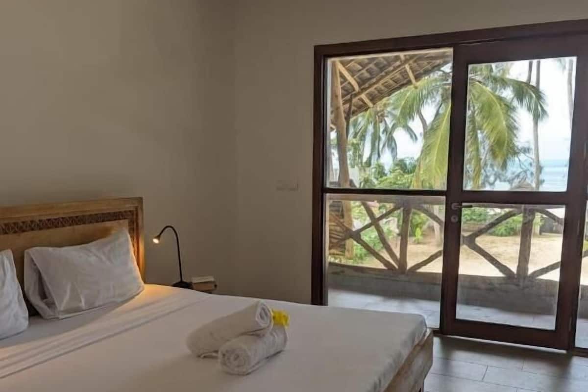 A single bed with white linens and a rolled towel, a wooden headboard, and a view of palm trees and a beach through a glass door.