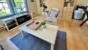 A living room with a TV, a white table, a wicker chair, a bookshelf, and a large window. - Sandy Toes Retreat (Ogunquit)