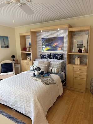 A bedroom with a bed, a headboard with artwork, a wooden wardrobe, and a chair.