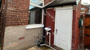 Een klein, afgesloten buitengebied met een witte deur, een raam met zonwering, en een witte wasbak met leidingen.