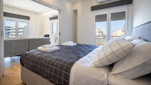 A modern bedroom with a large bed, a seating area, and a view of the city. - Attiki Square & Metro Modern 2-Bedroom Apt by MPS (Athina)