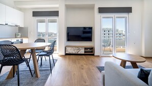 A modern living room with a dining area, a flat-screen TV, and a balcony with a city view. - Attiki Square & Metro Modern 2-Bedroom Apt by MPS (Athina)