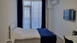 A hotel room with a bed, a desk with a chair, a TV mounted on the wall, and a window with curtains.