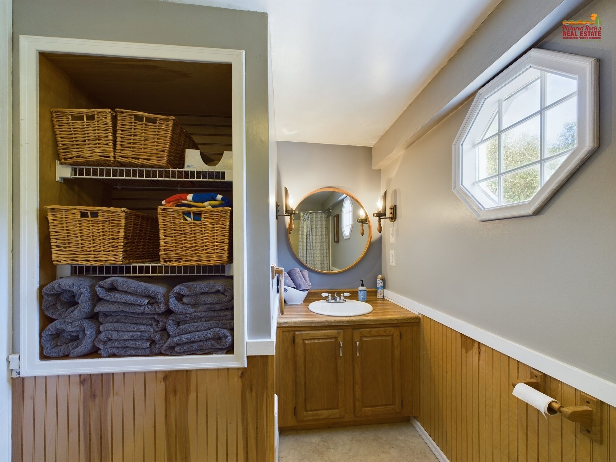 Ein Badezimmer mit einem Holzbadezimmerschrank, einem runden Spiegel, einem Fenster und einer Ablage mit Körben und Handtüchern.