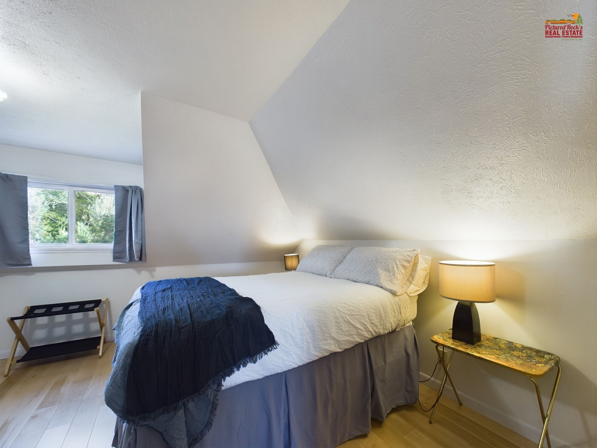 A bedroom with a bed, bedside lamps, a window with curtains, a small table, and a shelf.