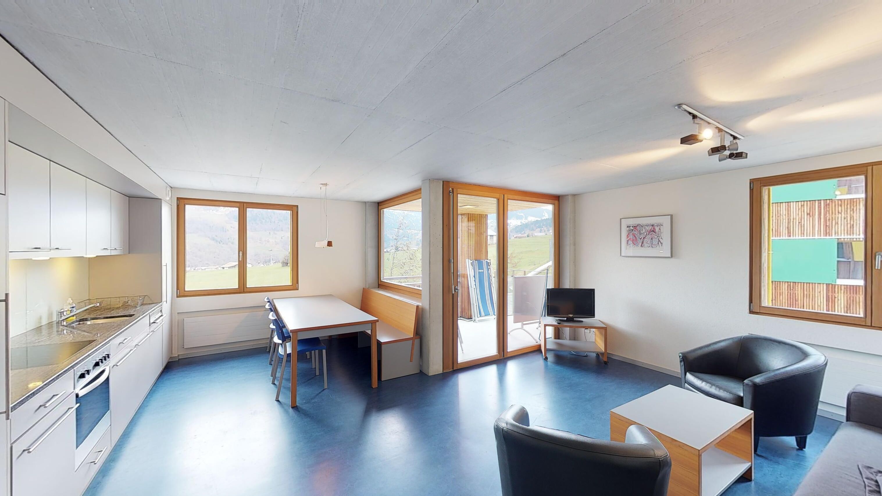 A compact living space with a kitchenette, a dining area, a seating area with a TV, and large windows offering views of the outdoors.