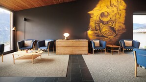 A modern lounge area with a wooden ceiling, a large wall mural, and a seating arrangement including sofas and a coffee table. - Reka-Feriendorf Disentis (Disentis)