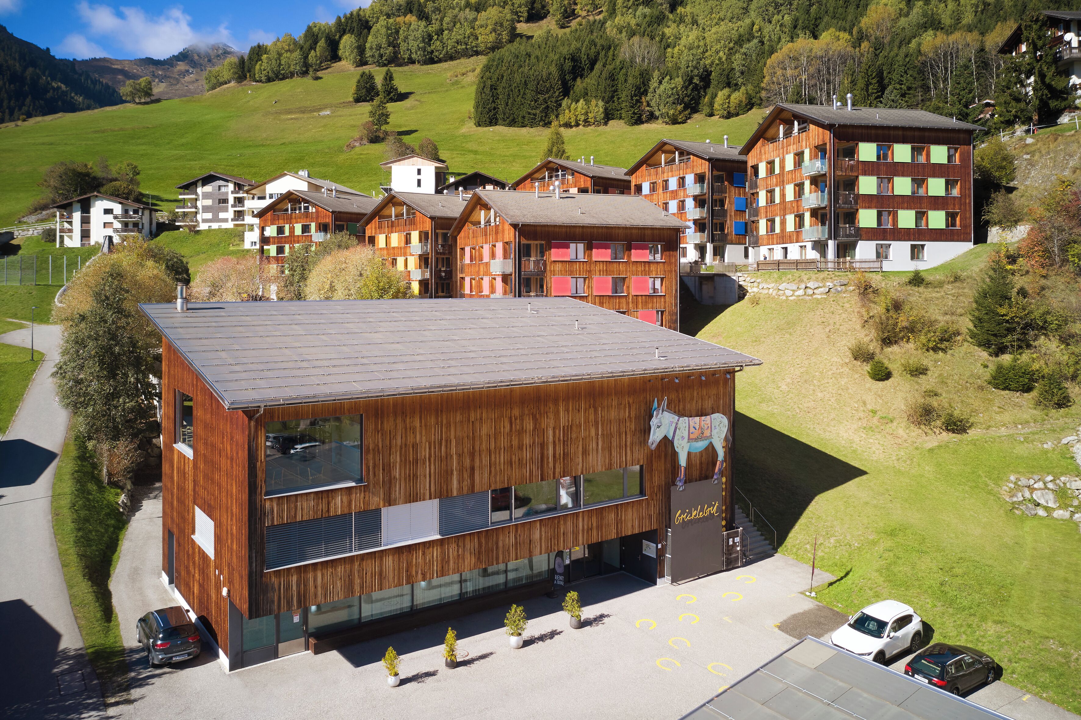 Ein modernes Gebäude mit großen Fenstern und einer Holzfassade, umgeben von einer grünen Landschaft und weiteren Gebäuden.