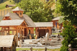 A rustic wooden building with a bell tower, a large wooden door, and a water wheel. - Reka-Feriendorf Disentis (Disentis)