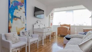 A modern living room with a white sofa, a white armchair, a wooden dining table, and a large abstract painting on the wall. - Barra da Tijuca 2 Bedroom Apartment on the Beach (Rio de Janeiro)