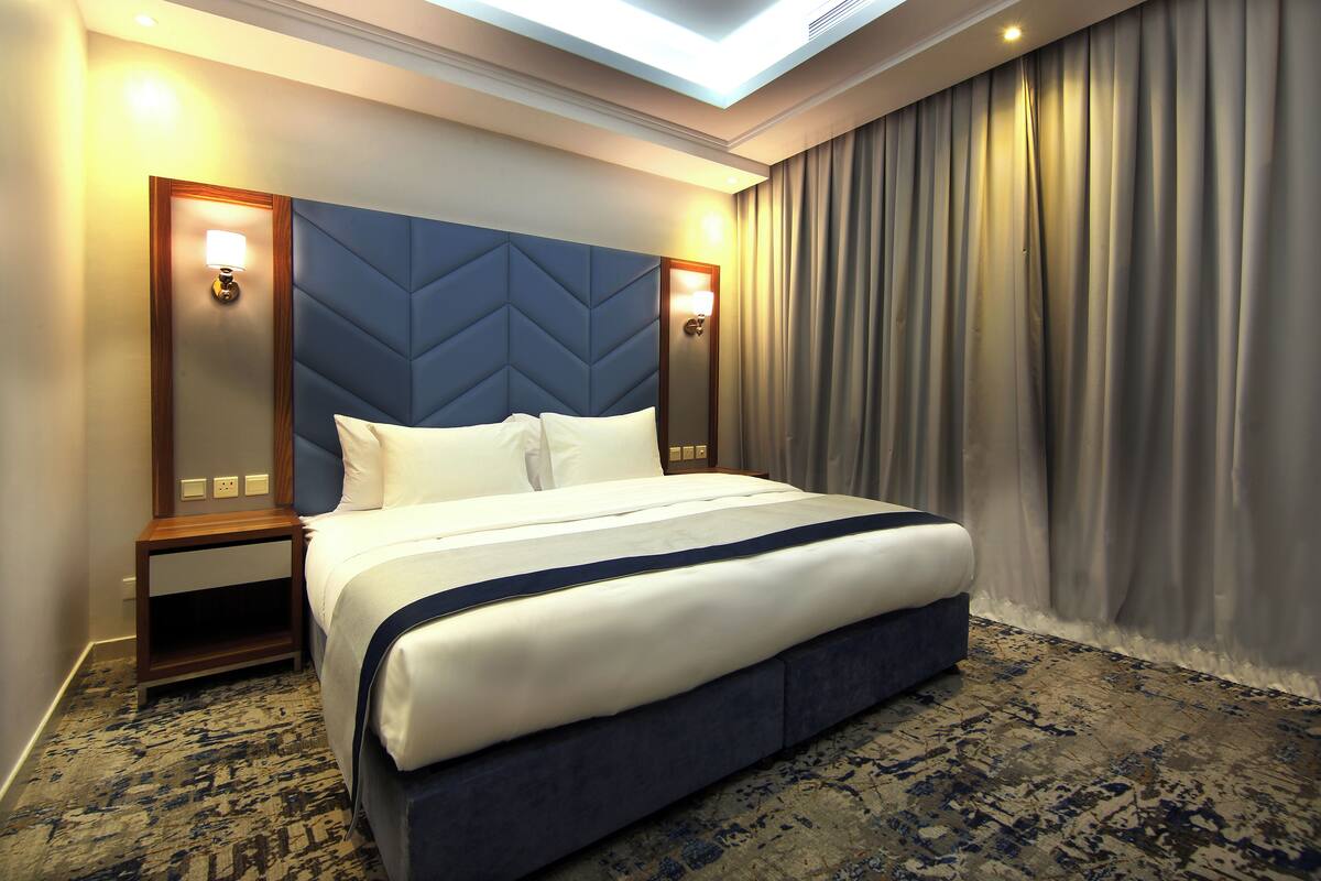 Een hotelkamer met een groot bed, een hoofdbord met een geometrisch patroon, twee nachtlampjes en een groot raam met gordijnen.