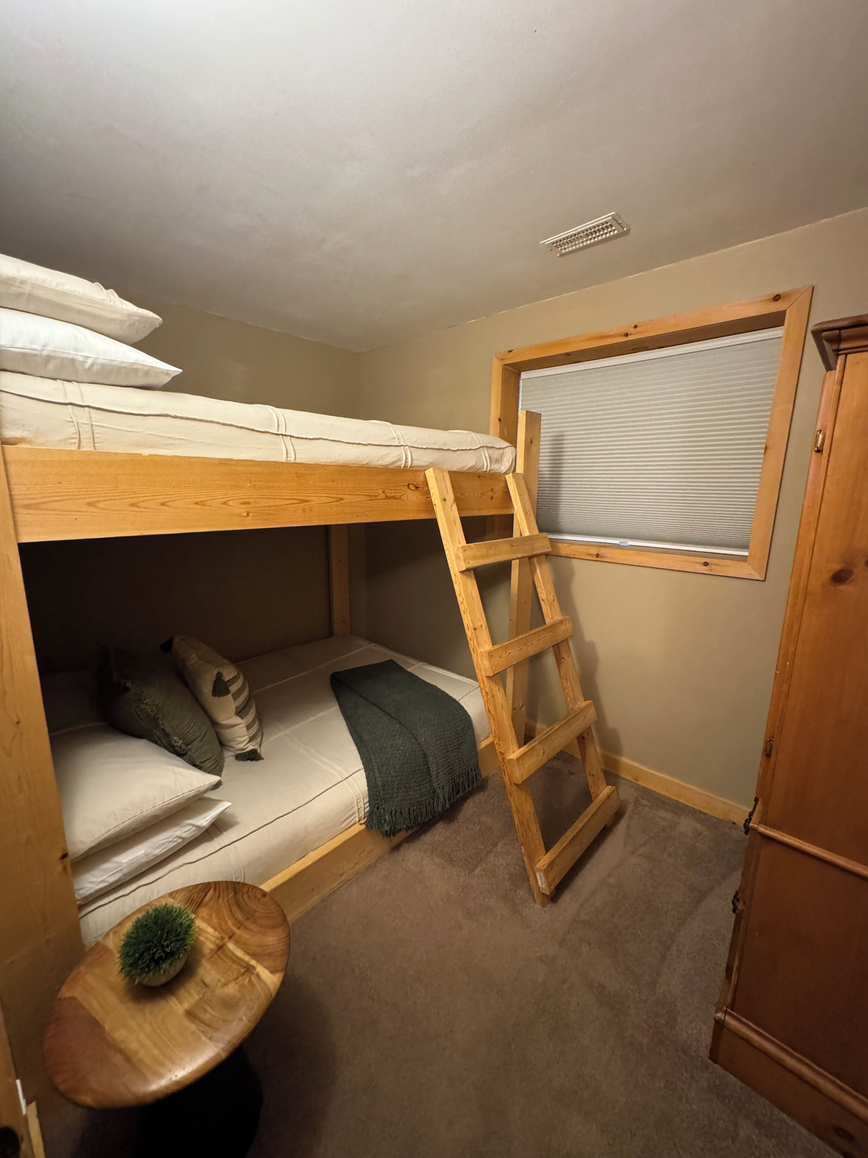 Un letto a castello con una scaletta in legno, un piccolo tavolino con una pianta e una finestra con persiane.