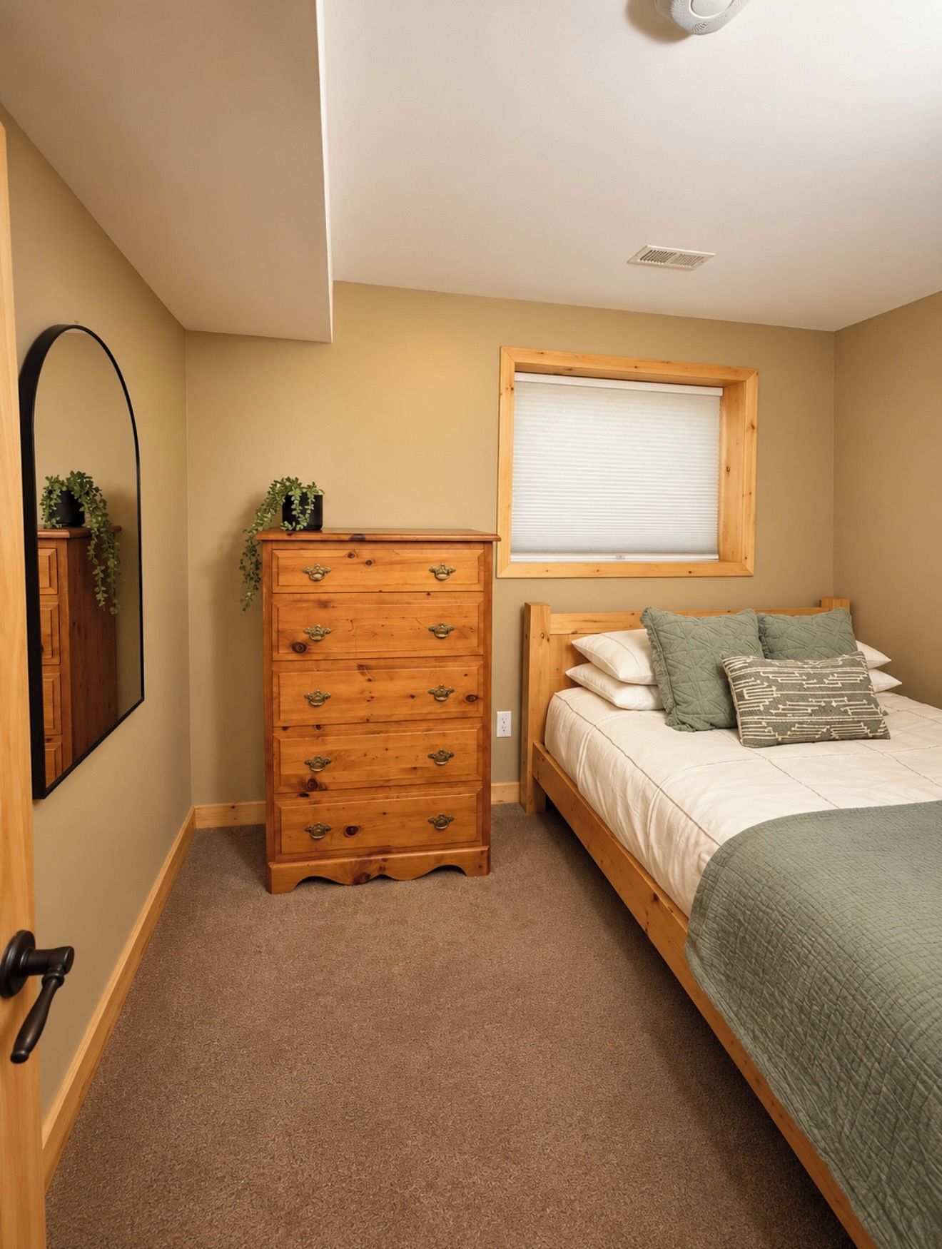 A bedroom with a wooden dresser, a bed with a green blanket, a window with blinds, and a ceiling light.