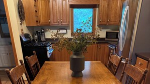 A wooden dining table with chairs, a vase with plants, and kitchen appliances in the background. - BirchView Trail *HOT TUB  (blue mountain)