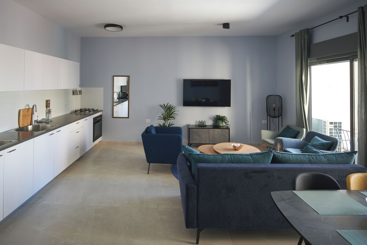 Une cuisine moderne avec des armoires blanches, une cuisinière à gaz et un évier. On y trouve un canapé bleu avec des coussins turquoise et une petite table ronde. Un téléviseur à écran plat est fixé au mur, et une plante est posée sur une table d’appoint.