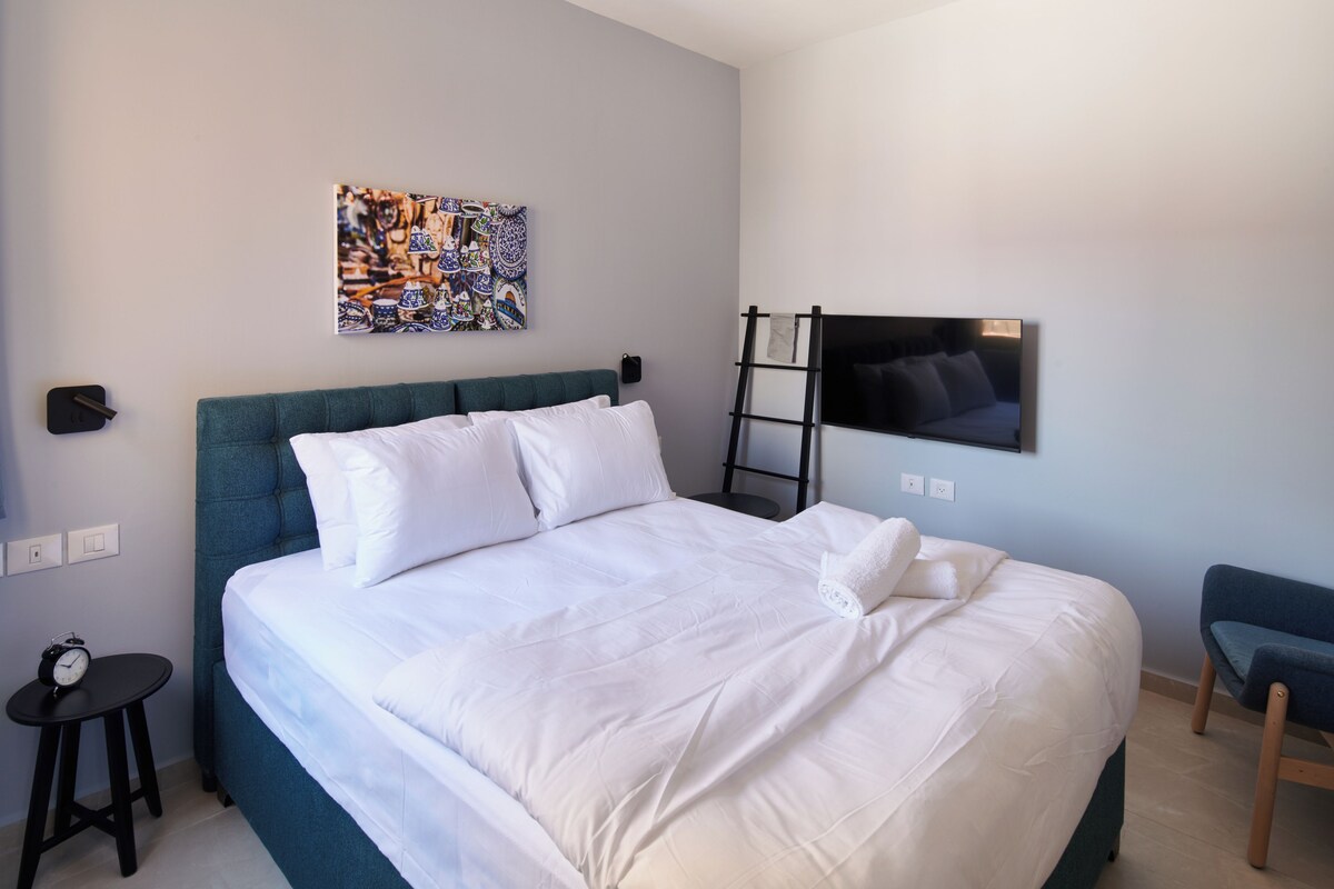 Una cama bien hecha con sábanas blancas, cabecera color turquesa y televisor montado en la pared.