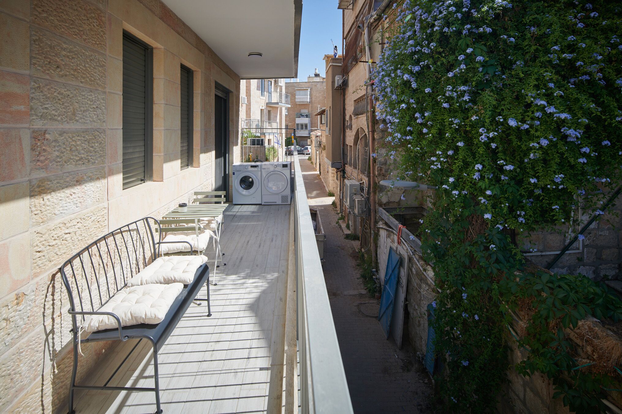 A narrow alleyway with a bench and a washing machine on the left, a wall with a climbing plant on the right, and buildings in the background.