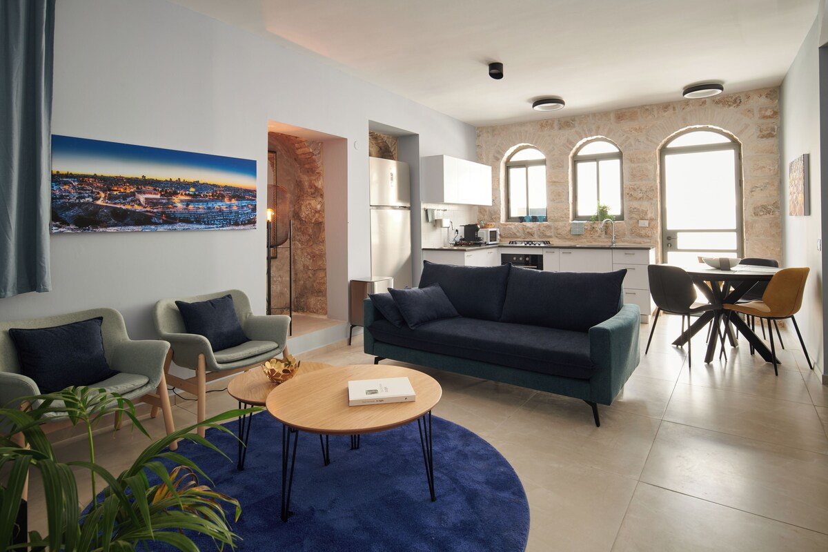 Een moderne woonkamer met een bank, fauteuils, een salontafel en een schilderij van een stadsgezicht.