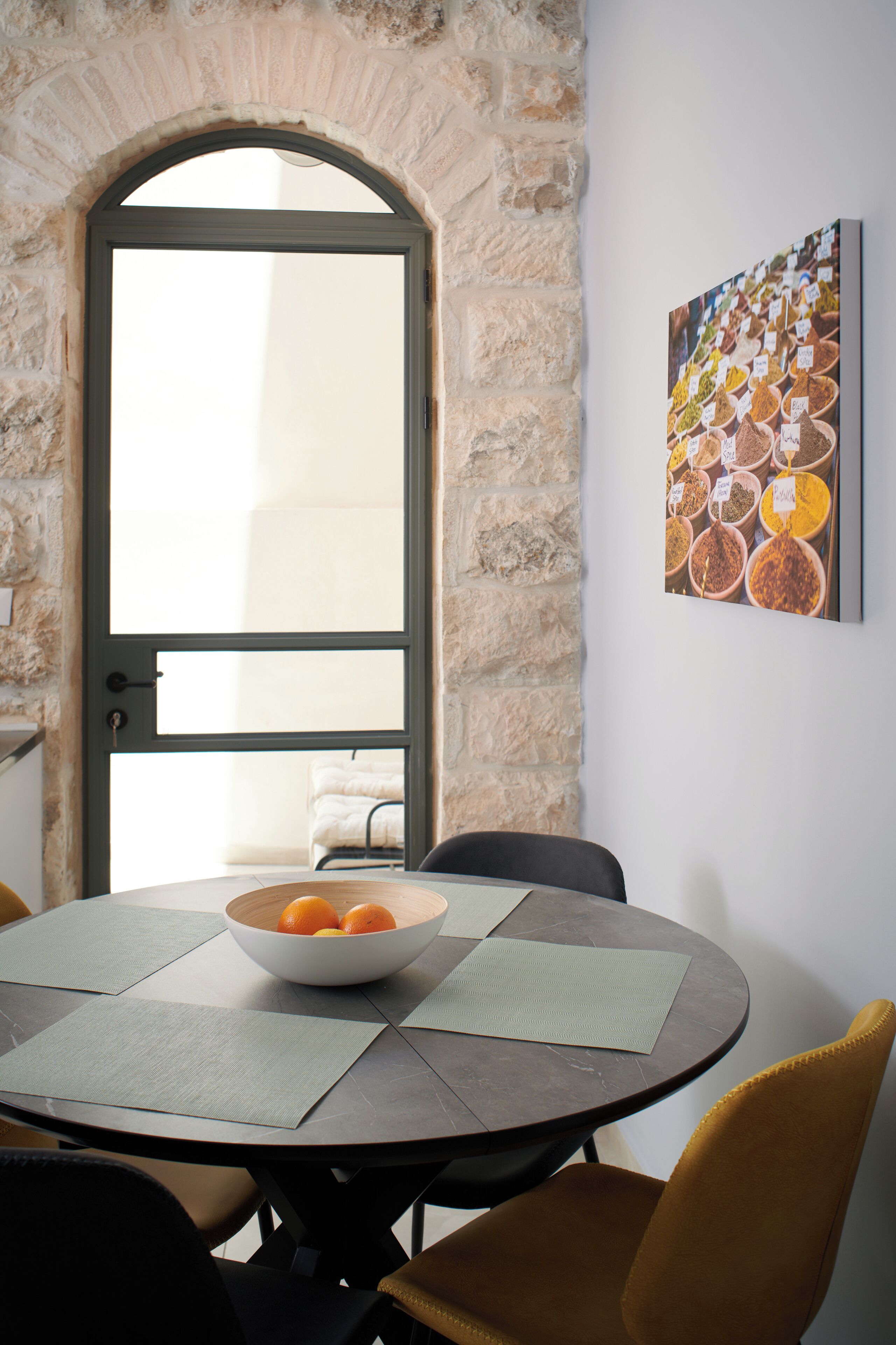 A dining area with a stone arch doorway, a round table with a bowl of oranges, and a spice-themed artwork on the wall.