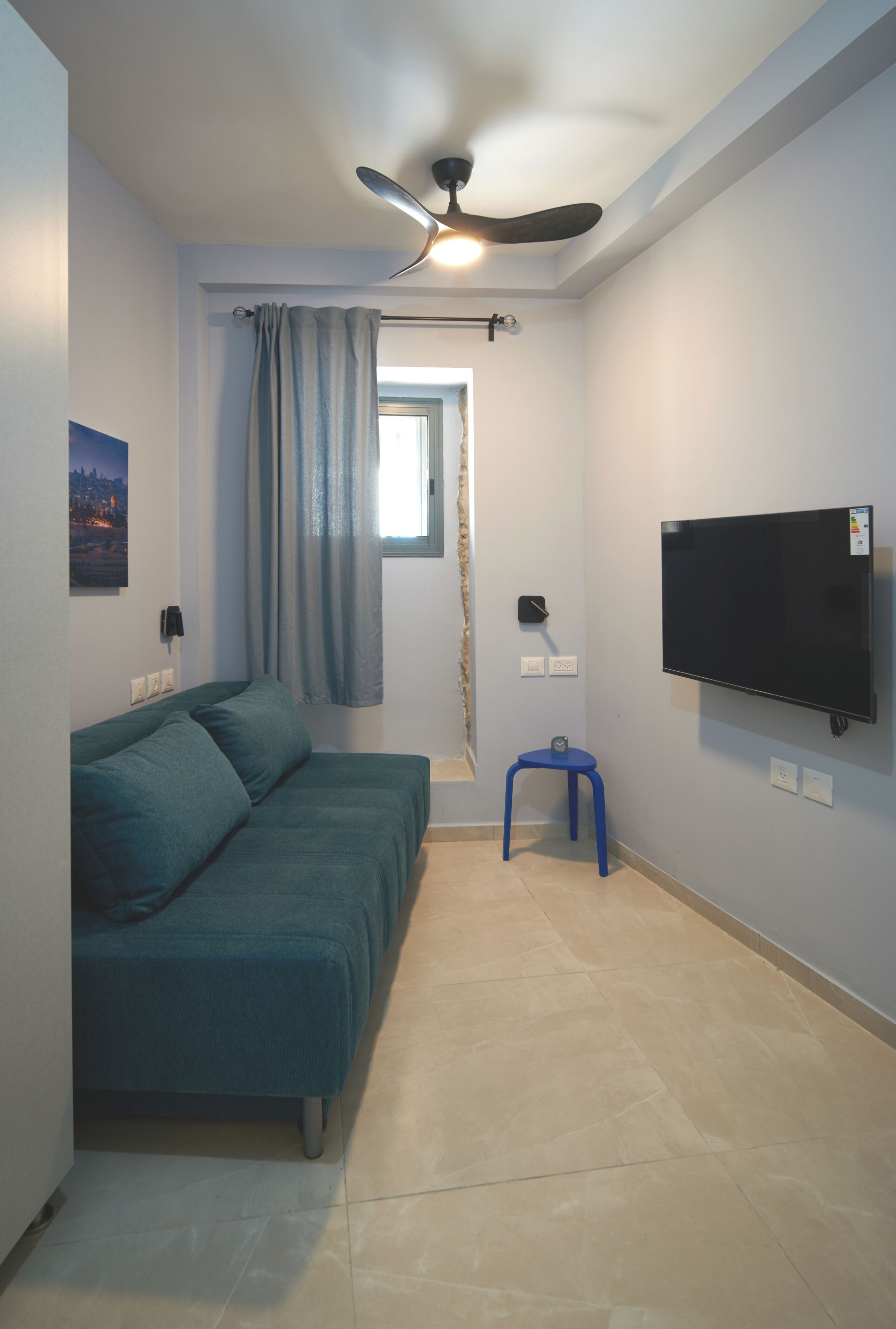 A small, modern room with a teal sofa, a ceiling fan, a TV mounted on the wall, and a small stool.