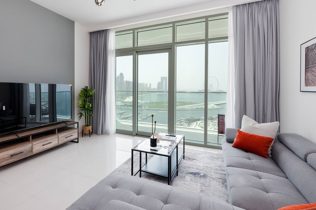 Una sala de estar moderna con un televisor grande, un sofá, una mesa de centro y vistas a un paisaje urbano y al agua.