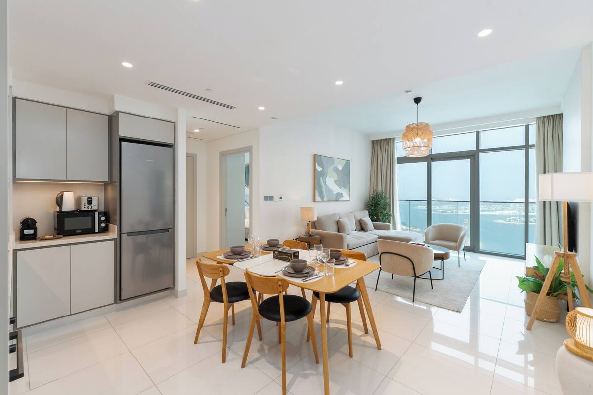 Una cocina moderna con microondas y refrigerador integrados, un comedor con mesa y sillas de madera, y una sala de estar con sofá y un amplio ventanal con vistas a la ciudad.