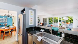 A modern kitchen with a stainless steel refrigerator, a black countertop, and a large window overlooking a garden. - 30+ Stay! Perfect Family Getaway!  W/Pool, Jacuzzi & Fire Pit (Palm Springs)