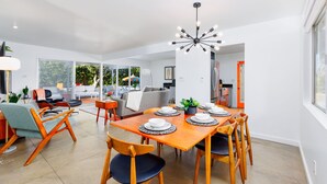 A modern dining area with a wooden table set for four, a contemporary chandelier, and a view of the outdoors through large windows. - 30+ Stay! Perfect Family Getaway!  W/Pool, Jacuzzi & Fire Pit (Palm Springs)