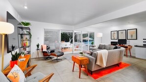 A modern living room with a sofa, chairs, a coffee table, and a TV. There is a bookshelf with plants and a desk with a lamp. - 30+ Stay! Perfect Family Getaway!  W/Pool, Jacuzzi & Fire Pit (Palm Springs)