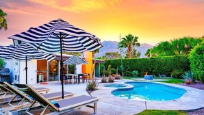 Un área junto a la piscina con sillones reclinables, una zona de estar techada con sombrillas y un jardín ajardinado.