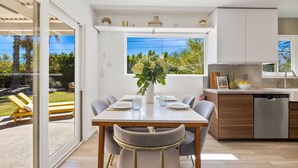 A modern kitchen with a built-in dishwasher, a dining area with a table set for four, and a large window overlooking a garden. - Sept Special Offer! 30+ Night Stay W/Pool/Hot Tub (Palm Springs)