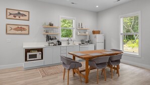 Cocina moderna con armarios blancos, una mesa de comedor de madera y sillas grises. Hay dos ventanas con persianas, un microondas y un fregadero.