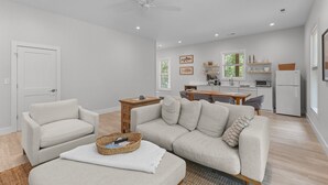 Un salón amplio con sofá, sillón y una pequeña mesa de madera. Se observa un ventilador de techo, y la habitación tiene una puerta y ventanas.