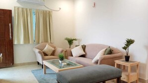 A living room with a sofa, armchair, ottoman, and a small side table with a plant. - Enkay Service Apartment (Hyderabad)