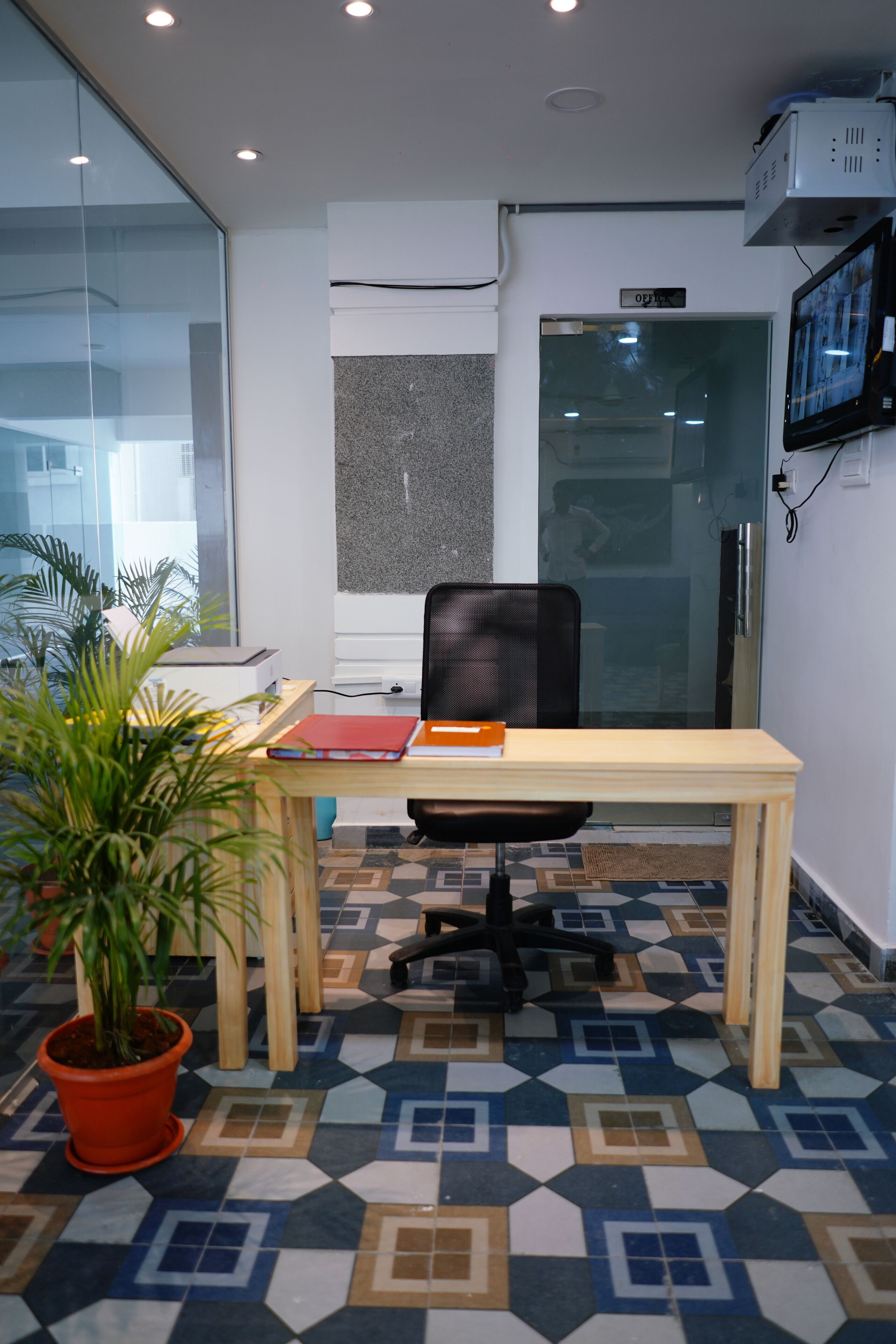 Ein modernes Büro mit Glastür, Holztisch, schwarzem Bürostuhl und einem Topf mit Pflanze.