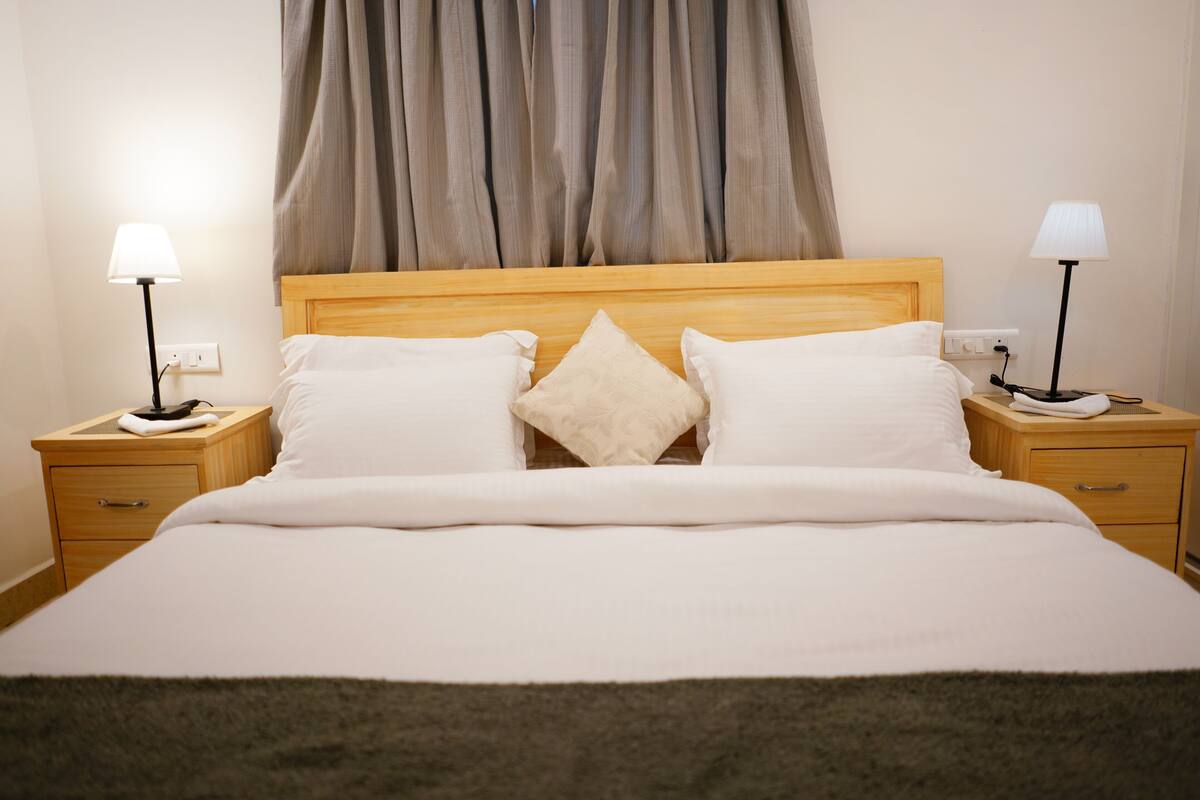 Una cama bien hecha con tablero de madera, dos mesitas de noche con lámparas, y un ventanal con cortinas.