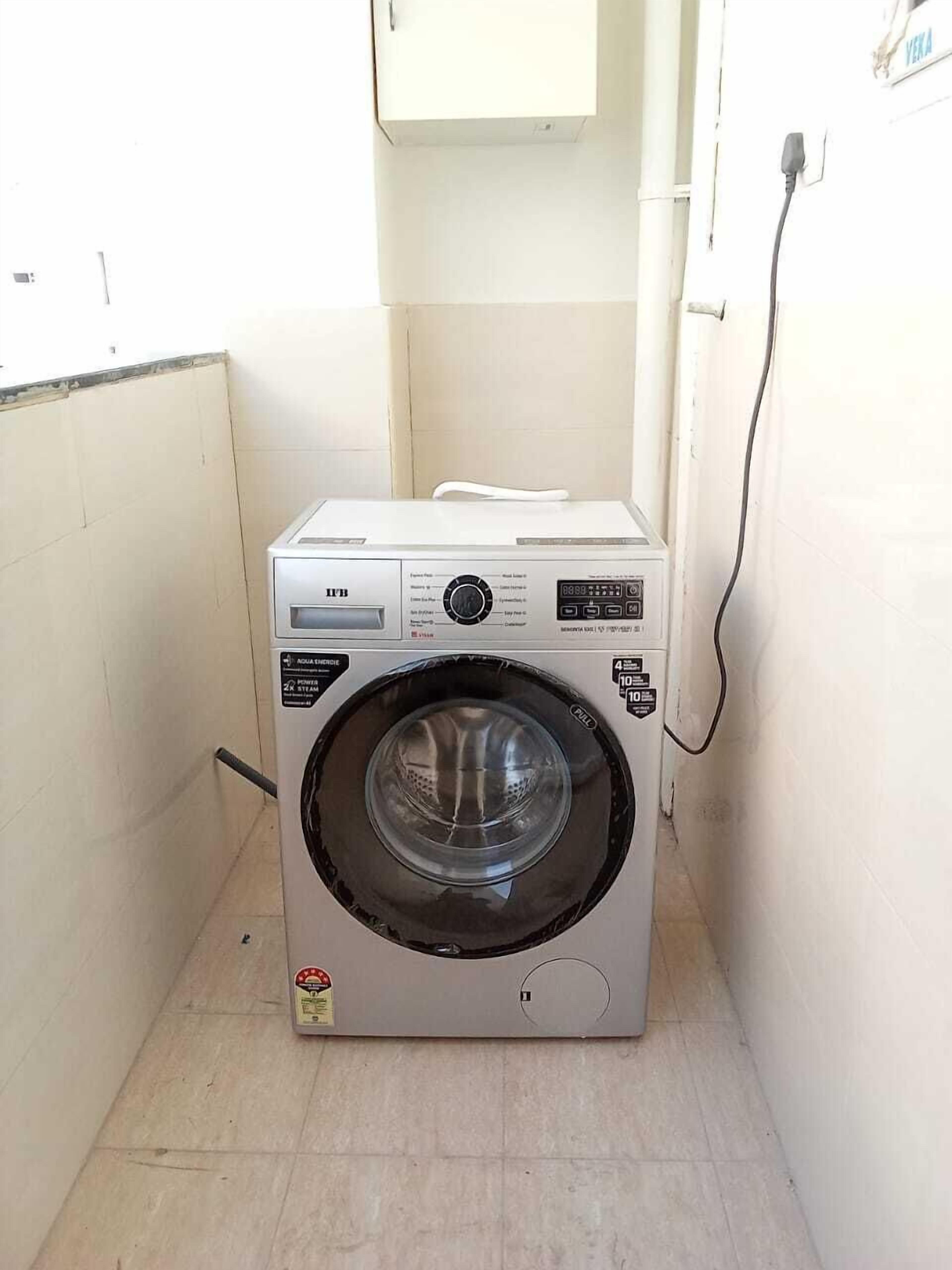 A compact, white front-loading washing machine in a small laundry room with tiled walls and floor.