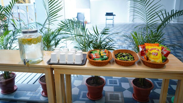 A table with a drink dispenser, cups, snacks, and potted plants. - Enkay Service Apartment (Hyderabad)
