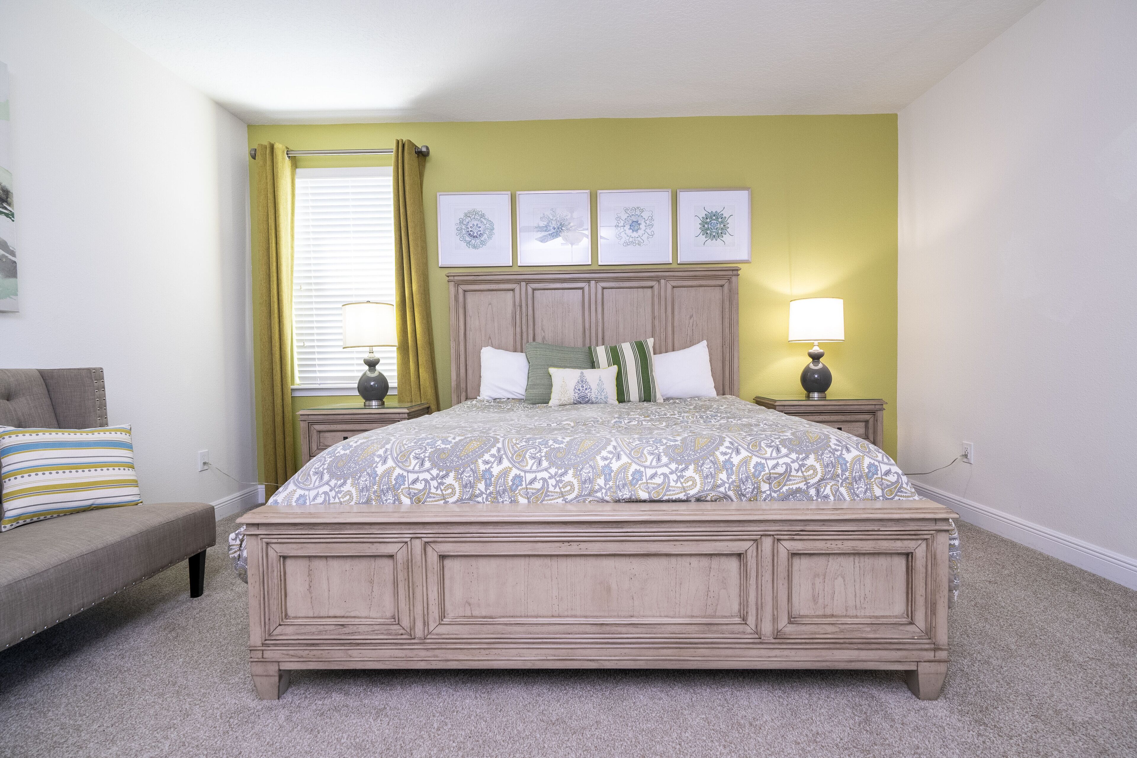 A bedroom with a bed, a nightstand, a lamp, a window with curtains, and a wall with framed artwork.