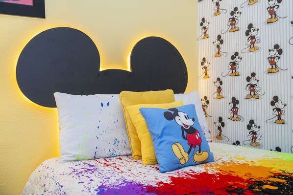 Una cama con almohadas y decoración temática de Mickey Mouse, además de papel tapiz con Mickey Mouse.