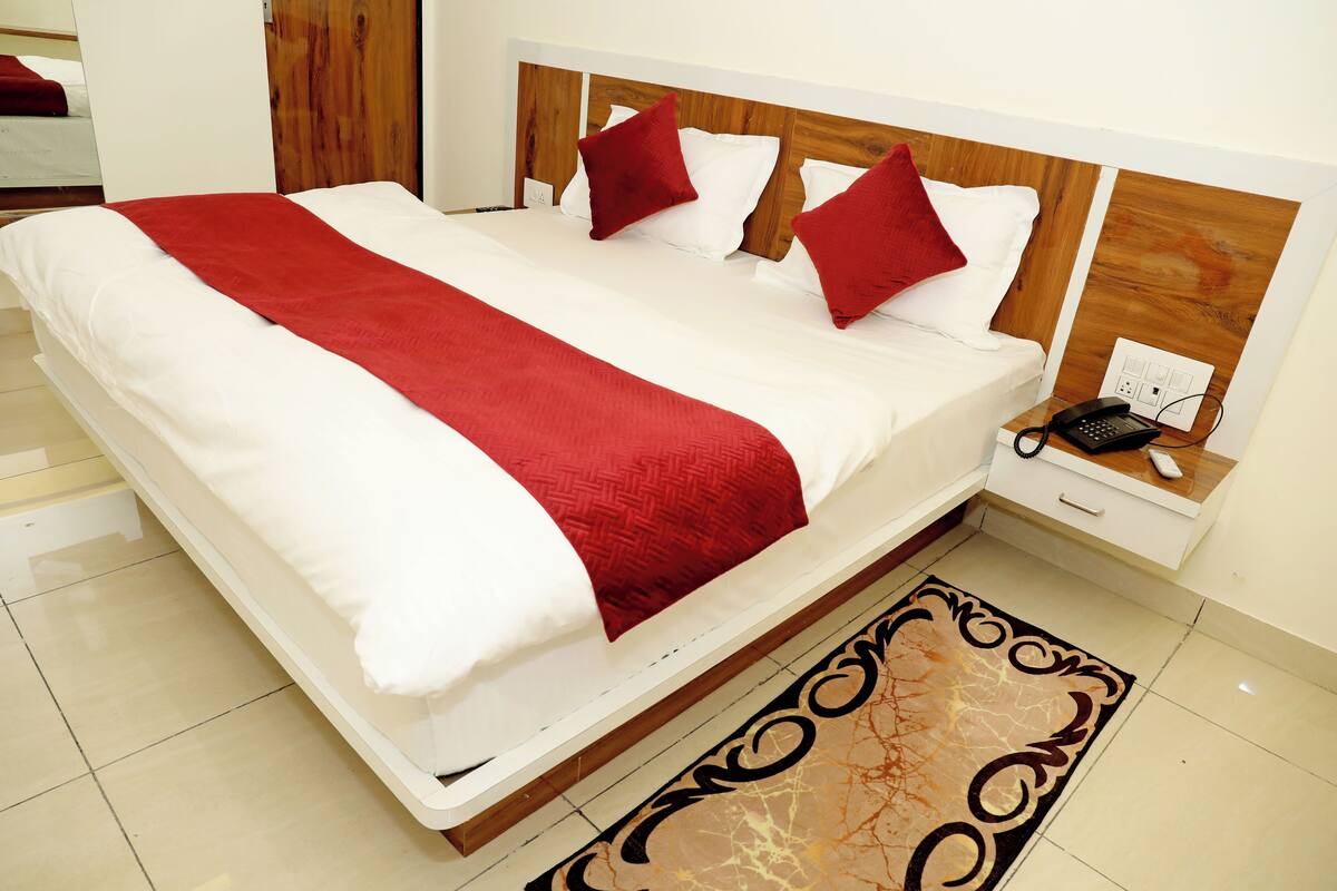 Un grand lit avec une tête de lit en bois, une literie blanche et des touches de couleur rouge. Il y a une table de chevet avec un téléphone et une prise électrique.