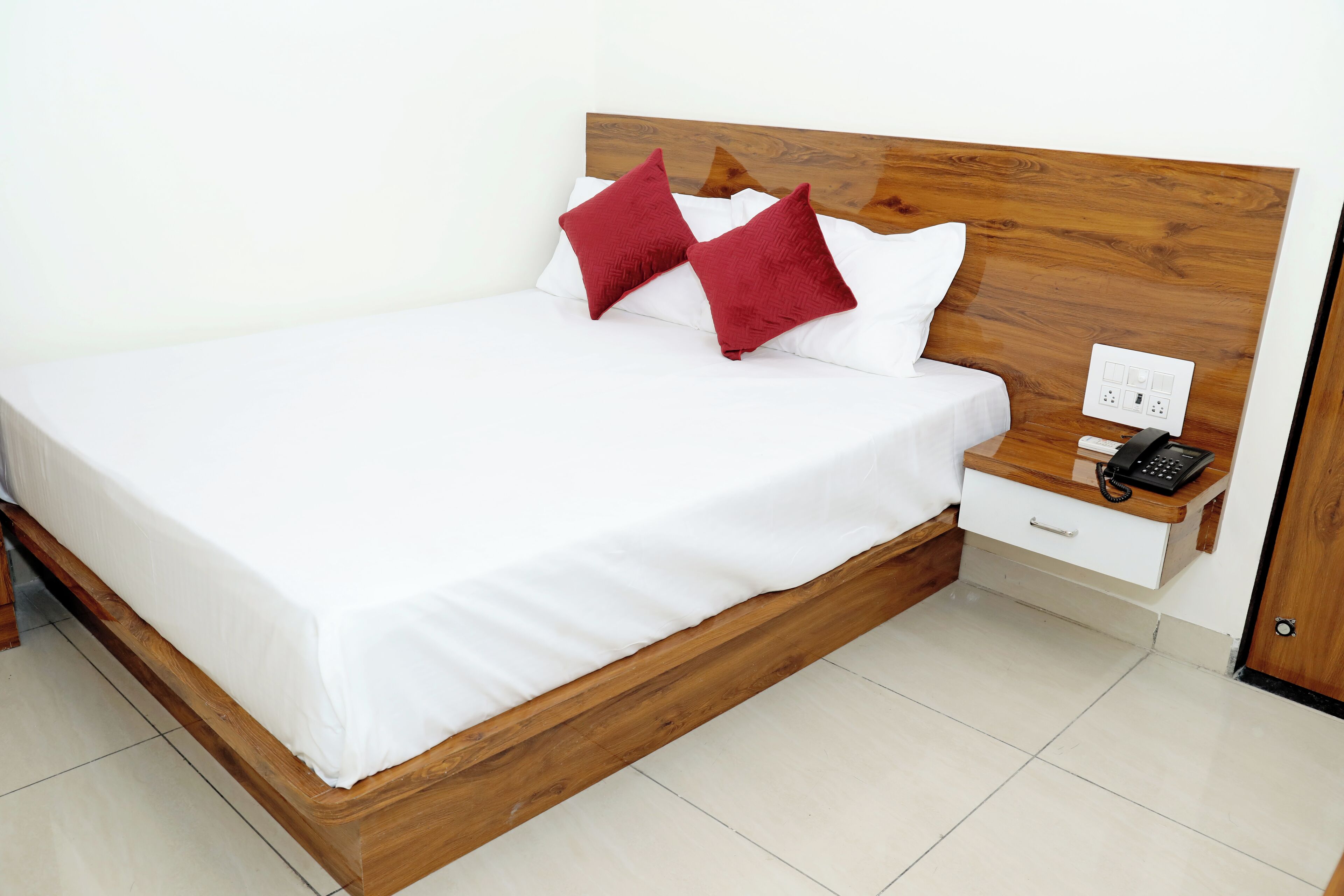 Una cama bien hecha con cabecera de madera, ropa de cama blanca y dos almohadas rojas. Se ve una mesita de noche con un teléfono y un enchufe.