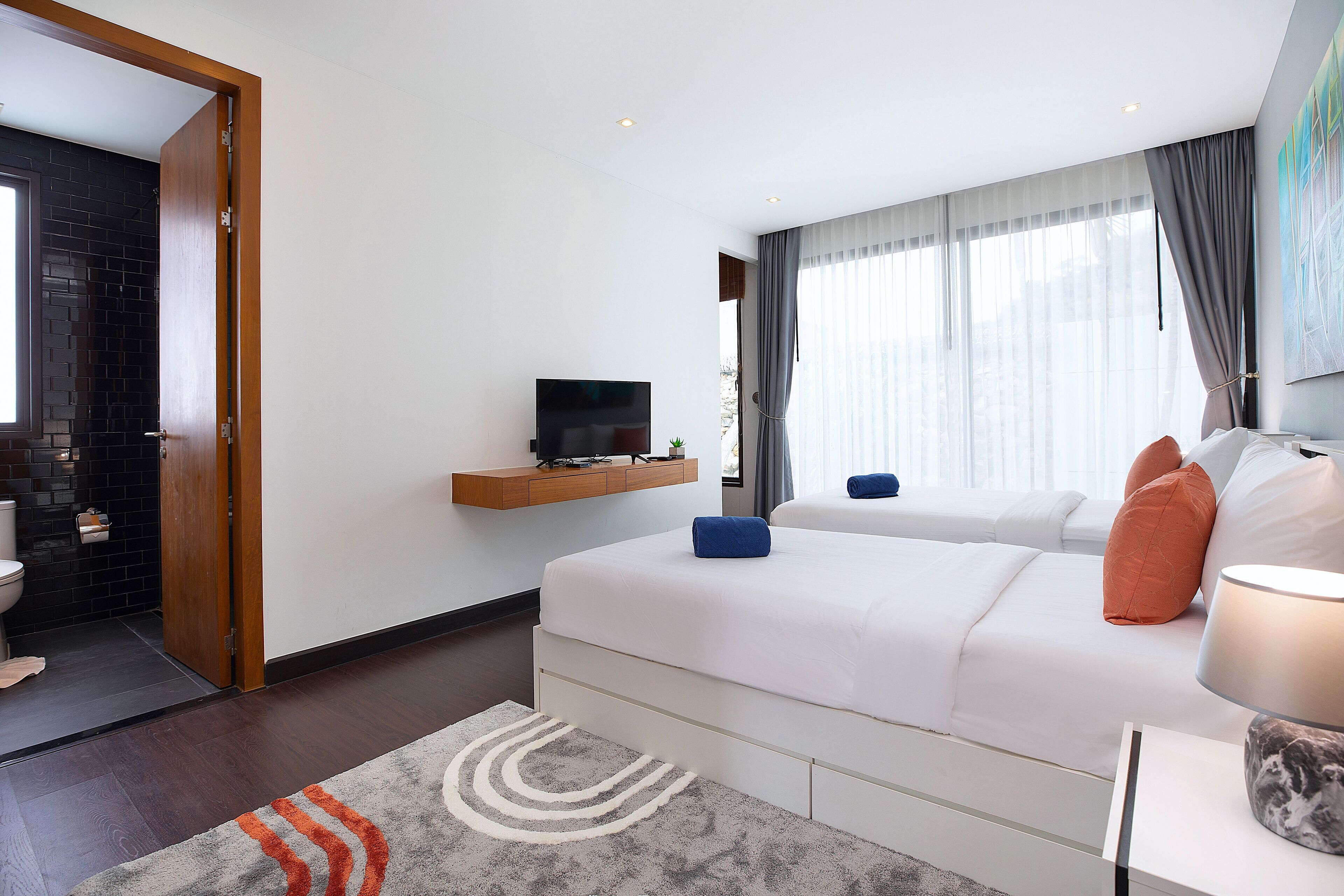 Habitación de hotel moderna con una cama grande, un televisor de pantalla plana montado en la pared, una mesita de noche con lámpara y vistas a la ciudad a través de amplios ventanales.