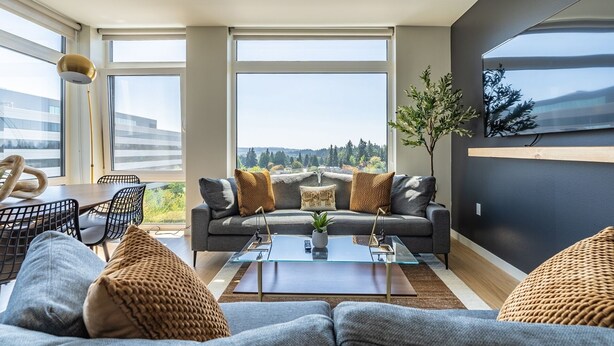 A modern living room with a sofa, coffee table, and a view of a forested area. - 2BR | Gym, Lounge | Bellevue | Sophari Downtown F (Bellevue)
