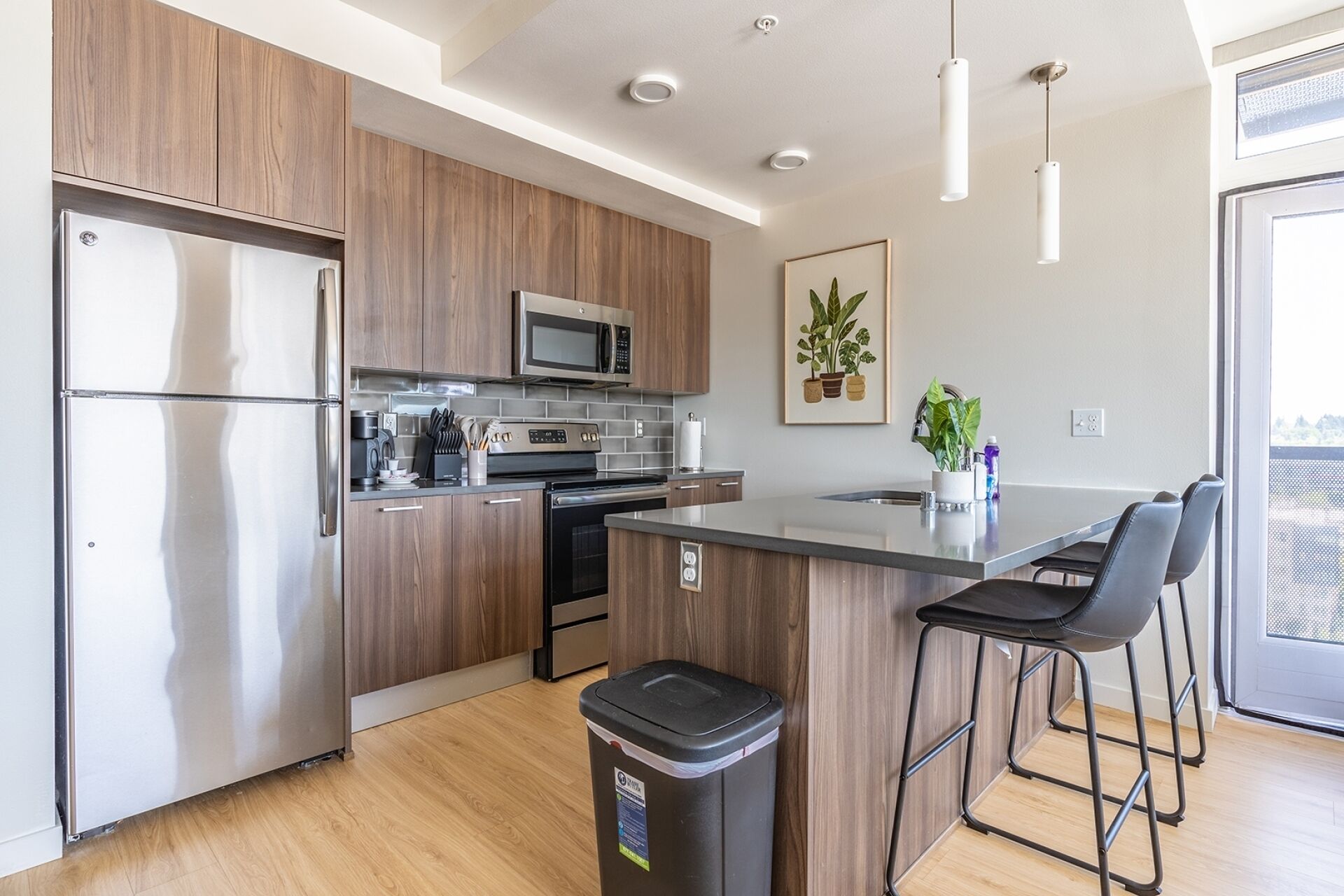 Una cocina moderna con armarios de madera, electrodomésticos de acero inoxidable, una isla central con taburetes altos y una planta en maceta.