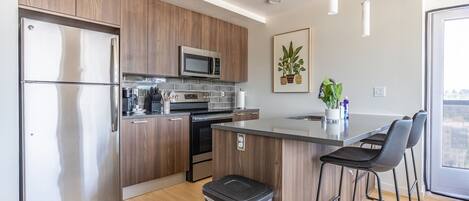 Una cocina moderna con armarios de madera, electrodomésticos de acero inoxidable, una isla central con taburetes altos y una planta en maceta.