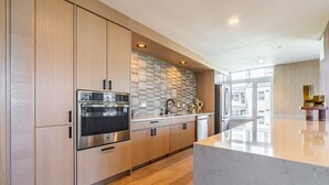 A modern kitchen with wooden cabinets, stainless steel appliances, a marble countertop, and a large window. - 2BR | Gym, Lounge | Bellevue | Sophari Downtown F (Bellevue)