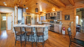 A rustic kitchen with a stone island, wooden bar stools, and a stone fireplace. - Dog Friendly Mountain Retreat w/ Bar & Hot Tub (McHenry)