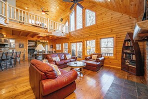 A spacious log cabin interior with a high ceiling, a balcony, leather sofas, a dining area, and a fireplace. - Dog Friendly Mountain Retreat w/ Bar & Hot Tub (McHenry)
