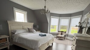 A bedroom with a large bed, a dresser, a chair, and a view of the outdoors through windows. - Charming Farmhouse on 2 acres, Sleeps 8. Only 5 mins from Richards Point beach (Borden-Carleton)